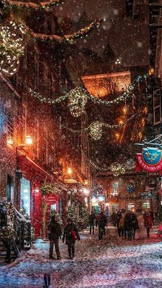 people walking down a snowy street at night with christmas lights on the buildings and trees