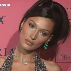 a woman with black hair and green earrings on her neck posing in front of a pink wall