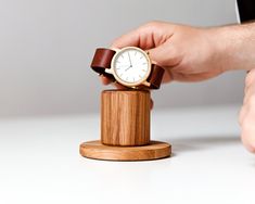 a person holding onto a wooden watch on top of a small wood stand with a white face and brown leather band