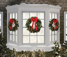 three christmas wreaths are hanging on the window sill in front of two evergreen trees