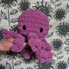 a hand holding a purple crocheted stuffed animal in front of a flowered background