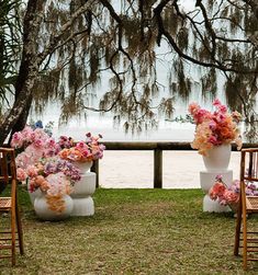 two chairs with flower arrangements on them in front of a tree and water behind them