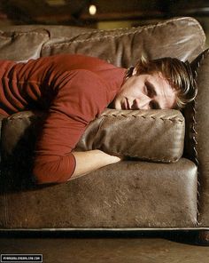 a man laying on top of a brown couch with his eyes closed and head resting on the pillow