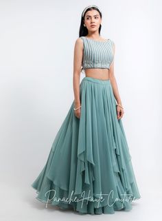 This exquisite ensemble features a stunning sage green tiered skirt crafted from lightweight georgette fabric, creating a flowy and airy silhouette. The skirt boasts multiple tiers that cascade gracefully down to the ankle, adding a touch of elegance to the overall look. Paired with the skirt is a beautiful blouse made from raw silk, adorned with intricate embroidery work using pearls, glass beads, and sequins petals. The embroidery adds a luxurious and glamorous touch to the blouse, making it p Georgette Skirt And Crop Top, Skirt And Crop Top Indian, Indowestern Dresses, Indowestern Gowns, Western Gowns, Indo Western Gown, Skirt And Crop Top, Crop Top Lehenga, Lehenga Saree Design