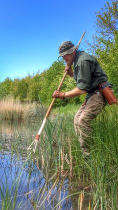 a man is standing in the water holding a stick and looking at some tall grass