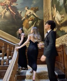 two women in black dresses are walking up the stairs to an ornate painting on the wall