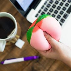 a person holding a small pink object in front of a laptop and cup of coffee