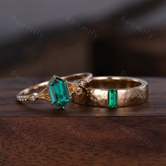 two gold wedding rings with emerald colored stones on top of each other, sitting on a wooden table