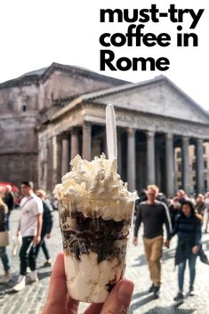 a hand holding an ice cream sundae in front of a building with people walking around