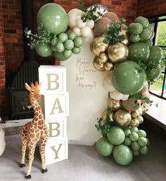 a giraffe standing next to a baby shower sign and balloon arch with greenery on it