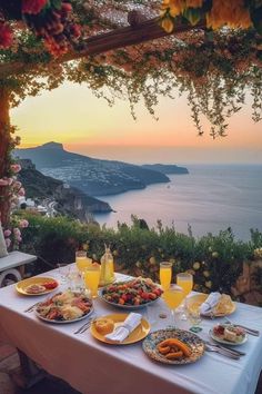 an outdoor dining table with food and drinks on it overlooking the ocean at sunset or sunrise