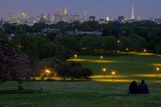 two people sitting on a bench in a park with the city lights lit up behind them