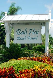 a sign for the beach resort in front of some trees and bushes with flowers around it