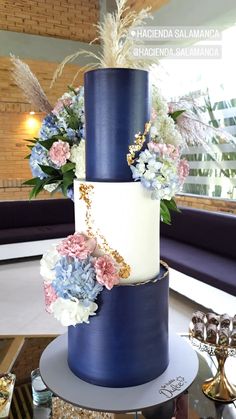 a three tiered blue and white wedding cake with flowers on the top, in front of a window