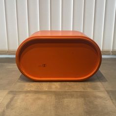 an orange bench sitting on top of a cement floor next to a white wall with vertical blinds