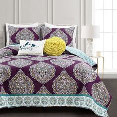 a purple and blue comforter set with pillows on the bed in front of a white wall