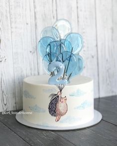 a white cake with blue balloons on top sitting on a wooden table next to a wall