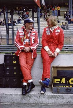 two men sitting next to each other on the side of a race track wearing red racing suits