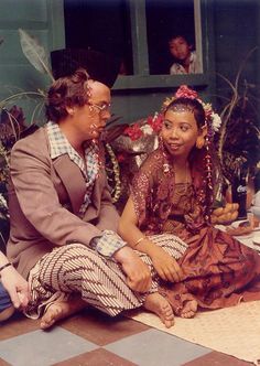 two people sitting on the ground talking to each other in front of some potted plants