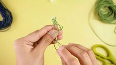 a person is working with yarn and scissors on a yellow surface, while another hand holds the string