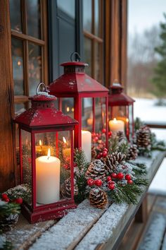 three red lanterns with lit candles sit on a window sill covered in snow and pine cones