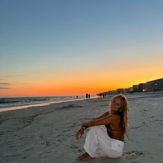 a woman sitting on top of a sandy beach next to the ocean at sun set