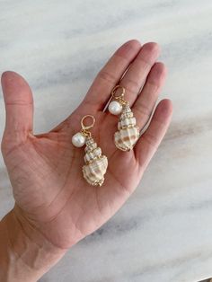 Beautifully crafted shell earrings with a pearl hanging, these lightweight earrings make a perfect addition to any jewelry collection. The pearl stands out against the natural shimmer of the gold painted shells, creating a stunning and eye-catching look. Shell-shaped Pearl Earrings With Pearl Charm, Gold Shell Drop Earrings, Ocean-inspired, Gold Pearl Drop Earrings With Ocean-inspired Style, Elegant Pearl Charm Earrings With Shell, Gold Ocean-inspired Shell Drop Earrings, Gold Mother Of Pearl Shell-shaped Earrings, Gold Shell Pearl Charm Earrings, Gold Shell Dangle Earrings, Pearl Shell With Pearl Charm