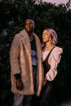 a man and woman standing next to each other in front of some bushes at sunset