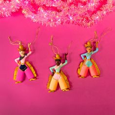 three toy figurines are hanging on a pink background with tinsel and lights