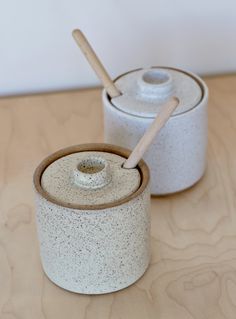 two white ceramic containers with wooden spoons in them on a wood table top next to each other