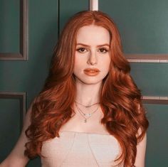 a woman with long red hair wearing a strapless top posing for a photo in front of a green wall