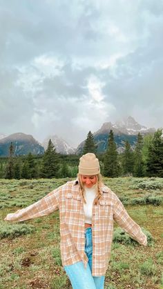 a woman in plaid shirt and blue jeans standing on grass with mountains in the background