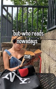 a woman sitting on top of a bench next to a bag and reading a book