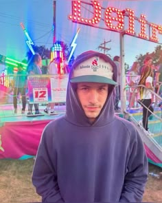 a man wearing a hoodie standing in front of a neon sign and carnival rides