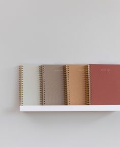 five notebooks lined up on top of each other in different colors and sizes, sitting on a white shelf