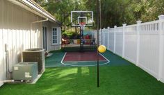 a backyard with artificial grass and a basketball court in the back yard, next to a white picket fence