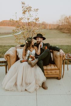 a bride and groom sitting on a couch in front of a tree with flowers around their necks