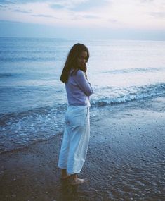 a woman standing on top of a sandy beach next to the ocean with her arms crossed