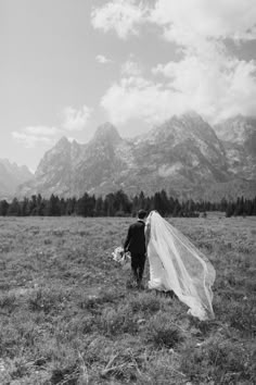 a bride and groom walking through the grass with mountains in the background