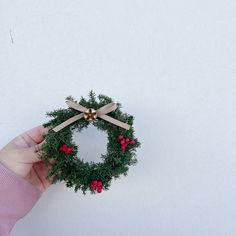 a hand holding a wreath with red berries and greenery on it against a white wall