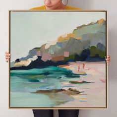 a woman holding up a large painting in front of her face, with the ocean and mountains behind her