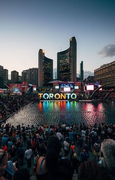 a large group of people standing around in front of a river with lights on it