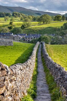 a stone wall and path leading to a village in the distance with green fields on either side