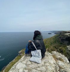 a woman sitting on top of a cliff next to the ocean with a bag in her hand