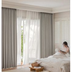 a woman sitting on top of a bed next to a window covered in white curtains