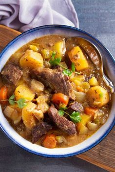 a bowl of stew with meat, potatoes and carrots on a wooden board next to a napkin