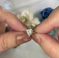 a woman's hands holding a ring with a heart shaped diamond on it and flowers in the background