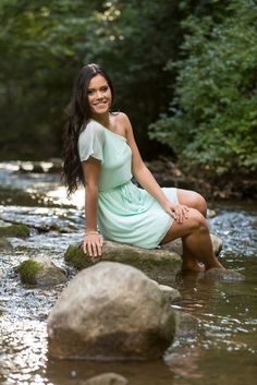 a beautiful young woman sitting on top of a rock next to a stream in the woods