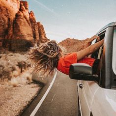 a woman leaning out the window of a car on a desert road with mountains in the background