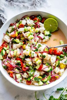 a white bowl filled with cucumber, red onion and tomato salad next to a lime wedge